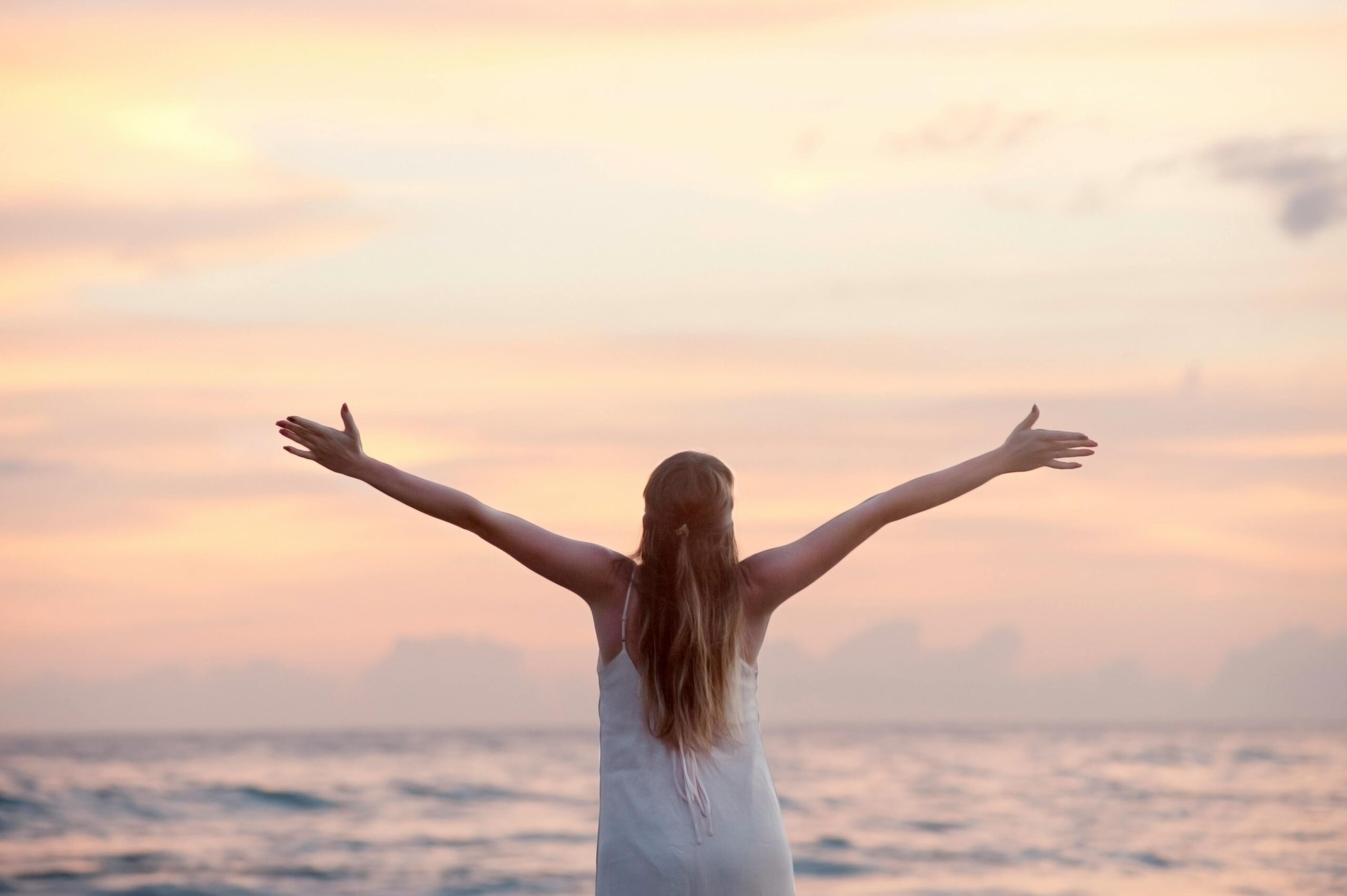woman standing with arms in the air while looking at the sun over the ocean