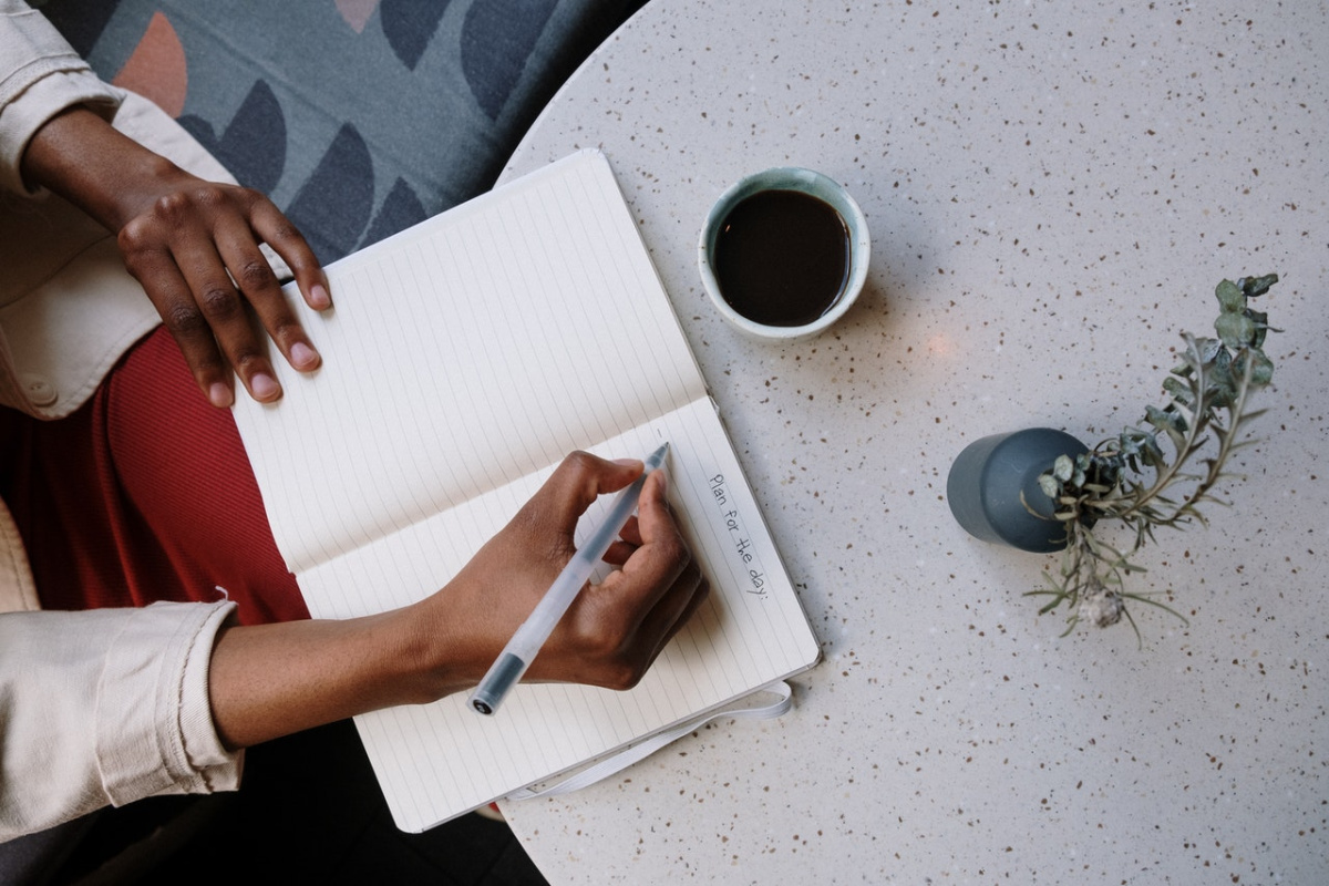 lady writing in a notebook with a cup of coffee and plant next to her