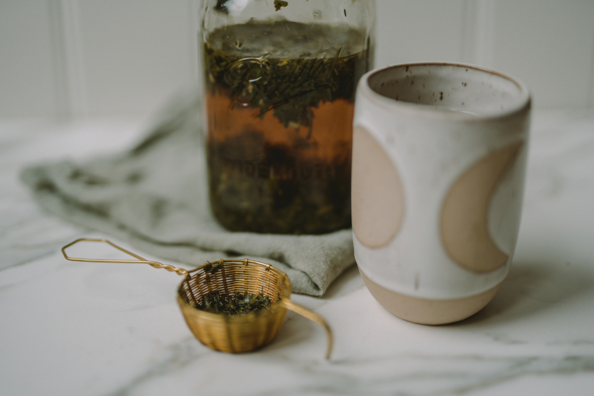 tea strainer with pregnancy tea herbs and jar of tea infusion with cup in the background