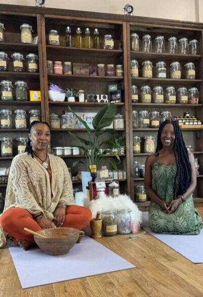 two women sitting on a floor with jars of herbs on bookshelf in background