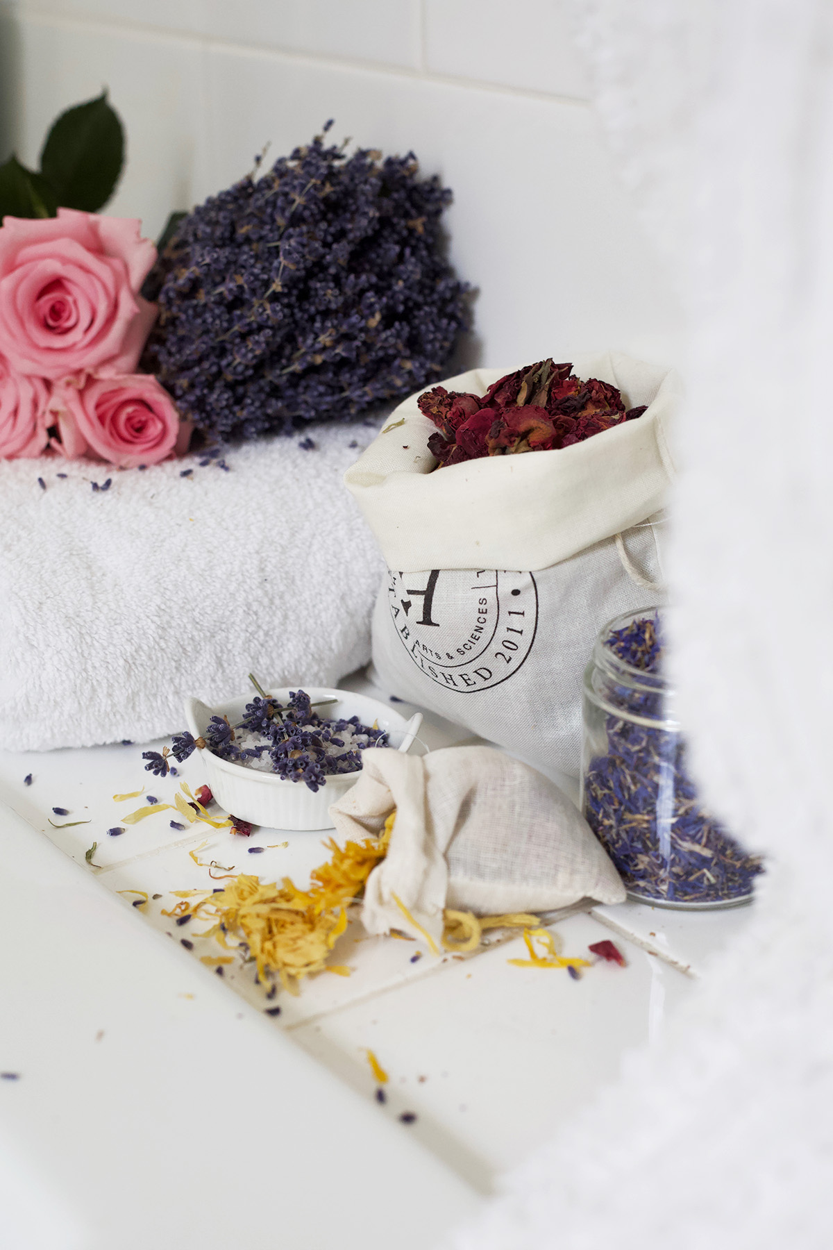 dried herbs sitting on a rolled up towel, in glass containers, and an Herbal Academy mulsin bag stuffed with dried rose petals