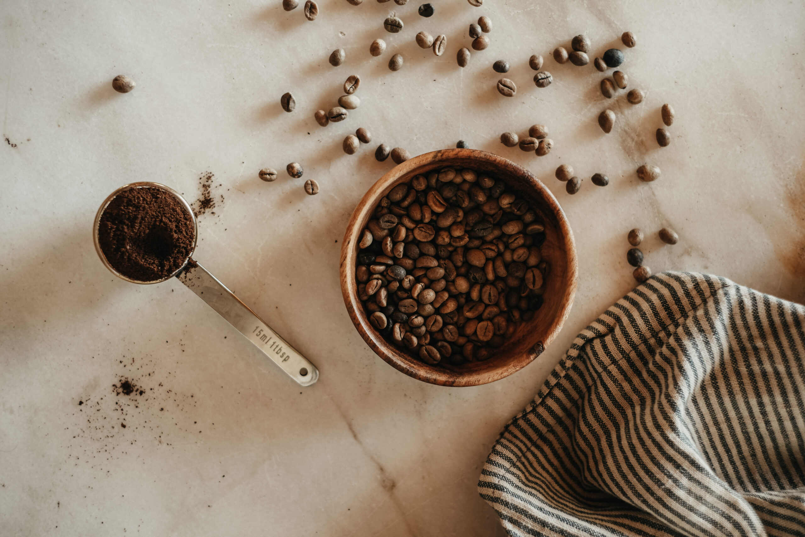 bowl of coffee beans with stainless steel measuring cup full of ground coffee and coffee beans scattered on the counter