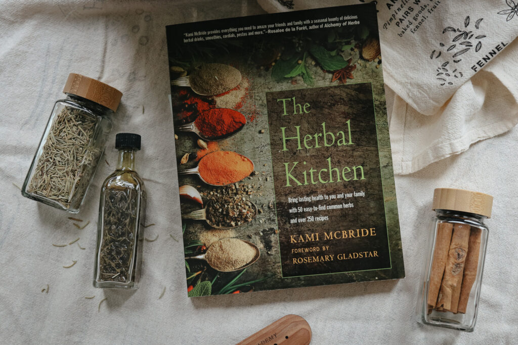 The Herbal Kitchen book on a tablecloth with bottles of herbs next to it