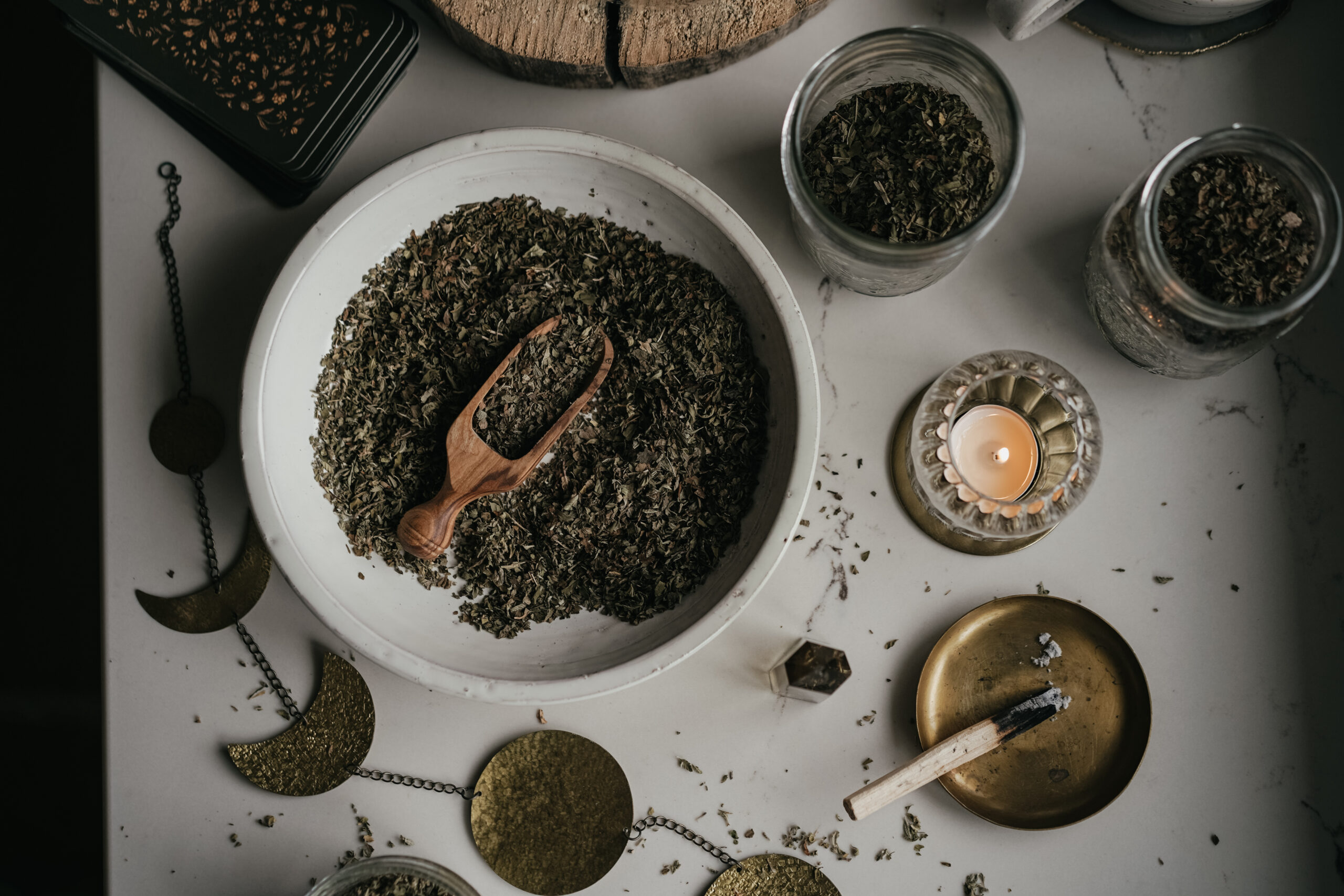 bowl of herbs and a wooden scoop with jars of dried herbs, a candle, and chain of moon phases on the table