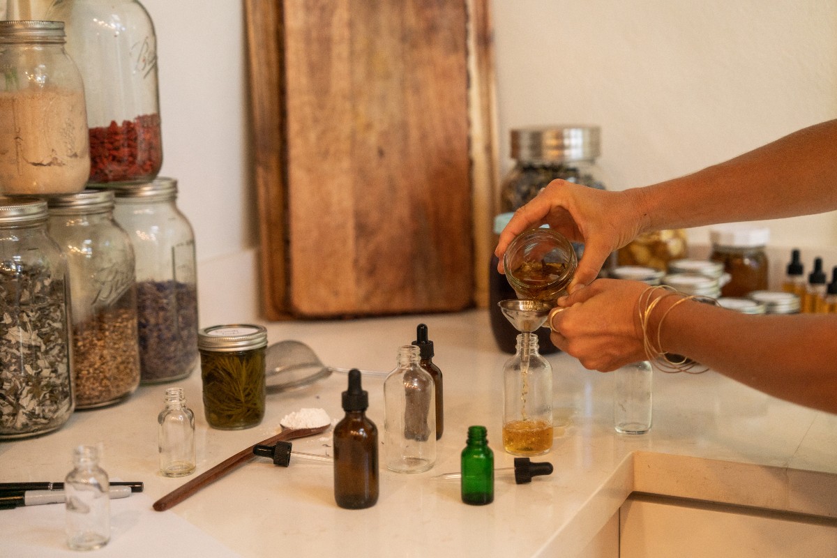 herbalist pouring infusion into a glass bottle with jars of herbs in the background