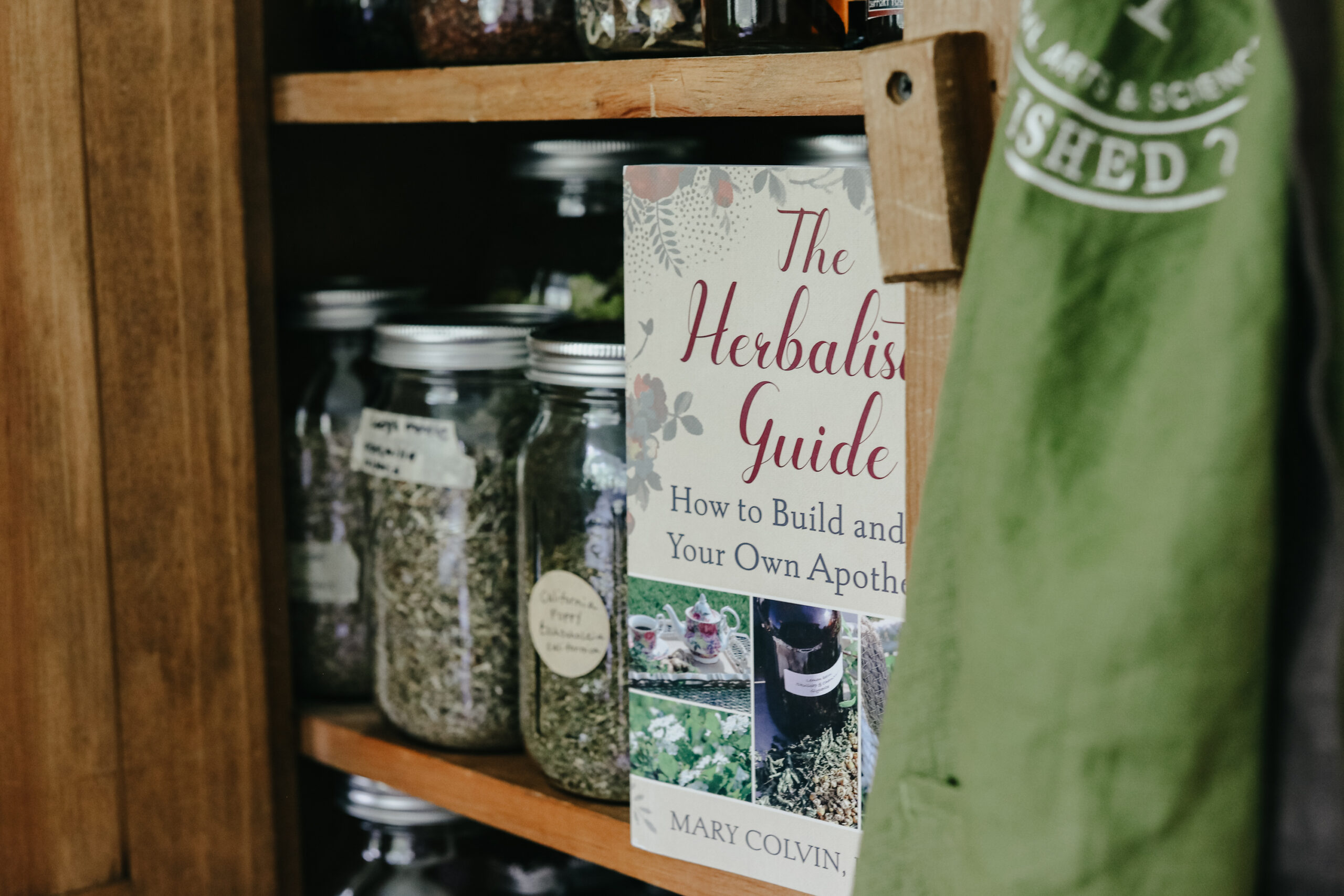 book and jars of herbs in a cabinet with apron hanging in the corner