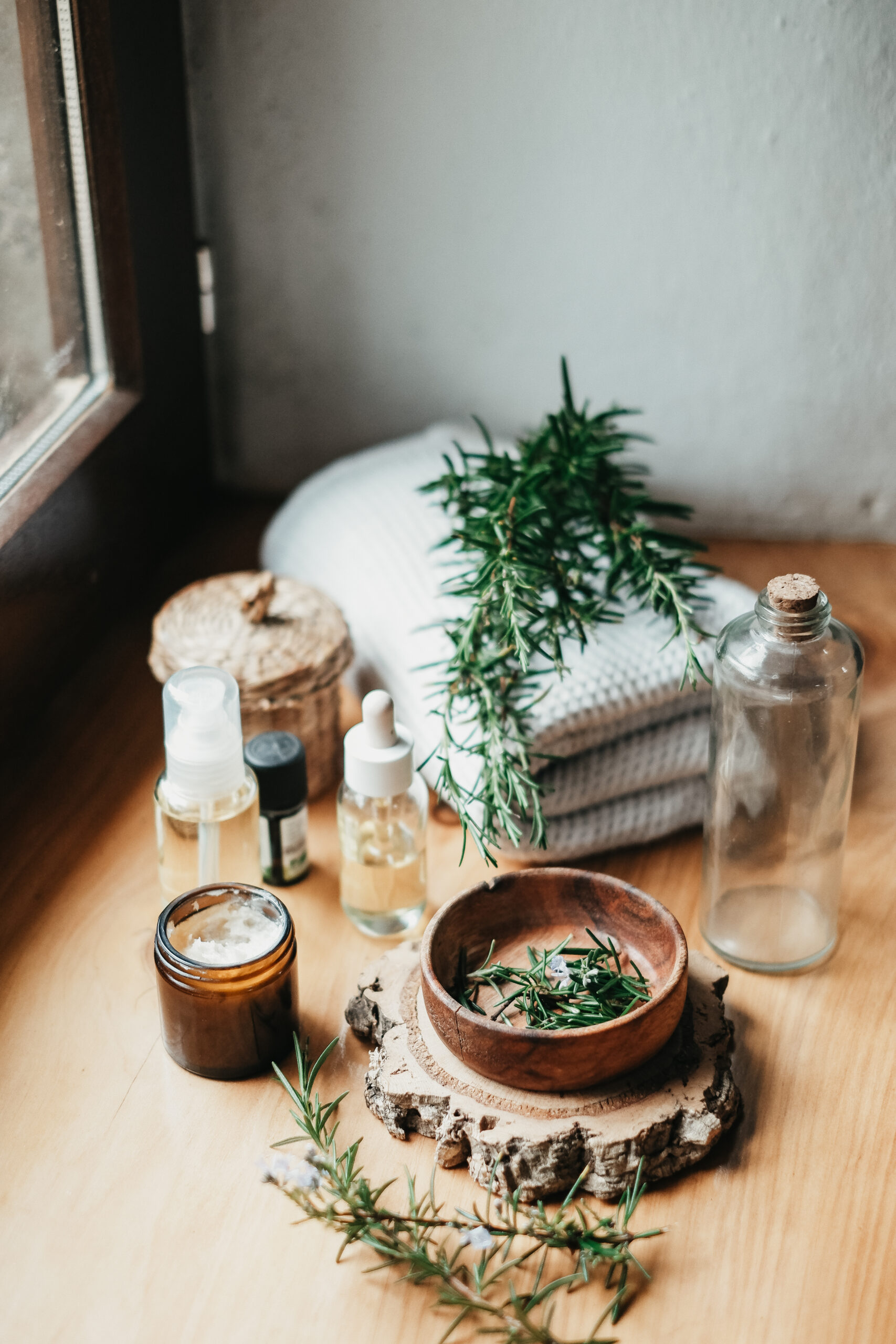 rosemary in a wooden bowl on a wood slab with blanket, herbs, and glass containers around it