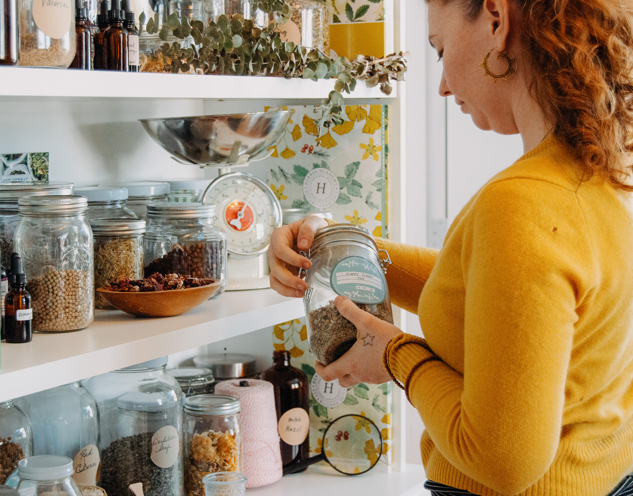 woman standing in front of shelves of jars of herbs while looking at a jar of herbs
