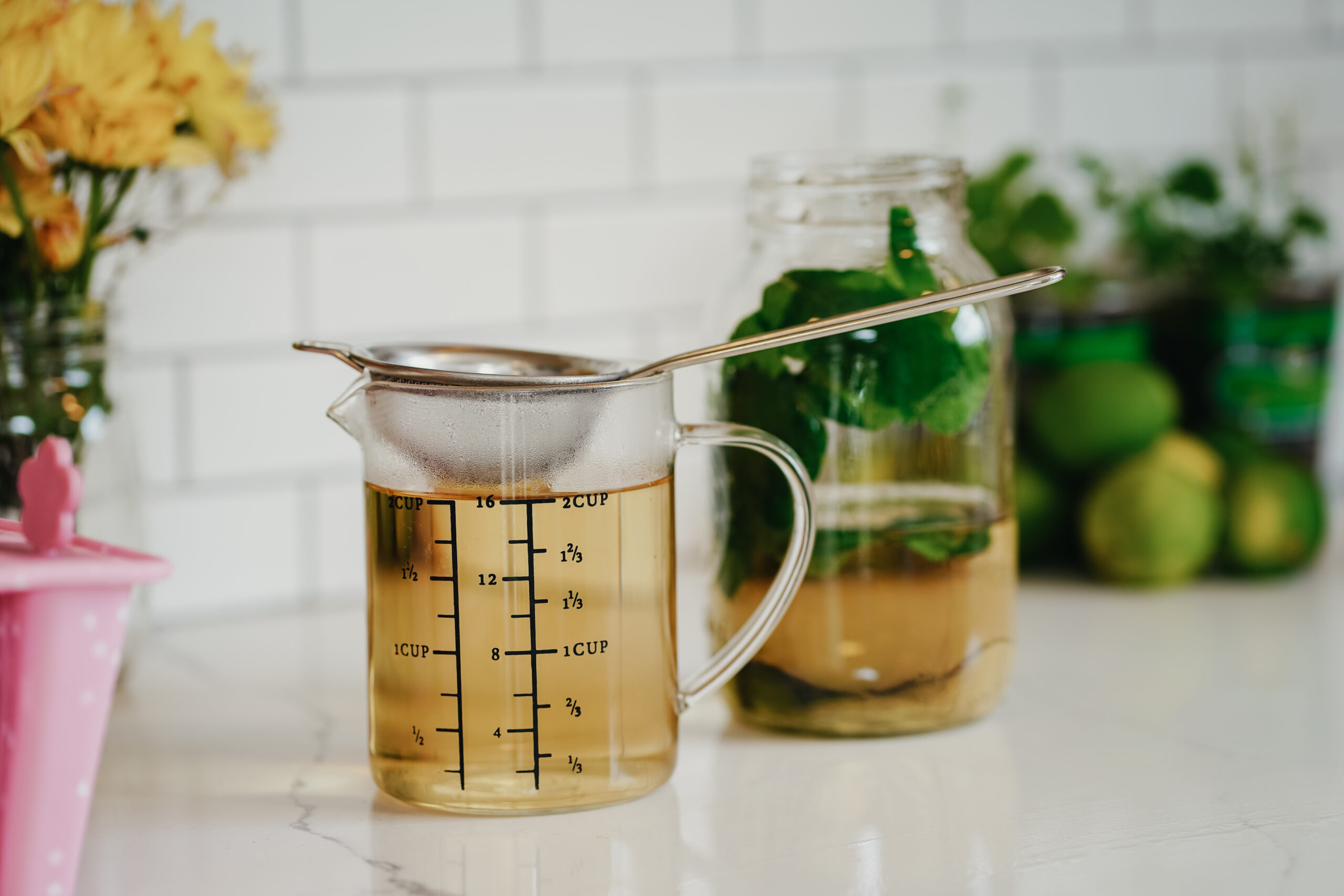 tea straining into a measuring cup with a jar of herbs in liquid in the background