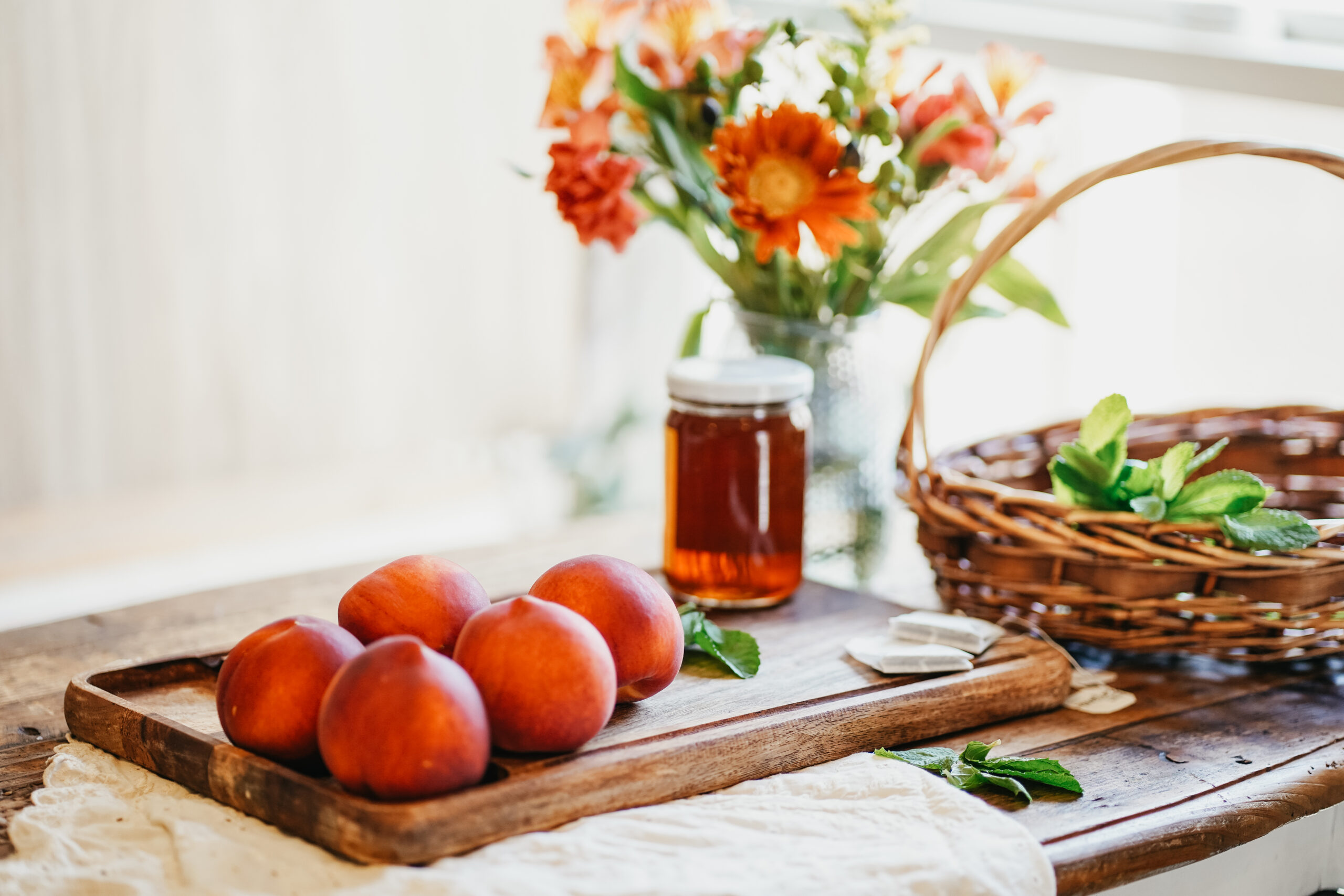 peaches and a jar of tea on a wood cutting board with a basket and vase of flowers in the background