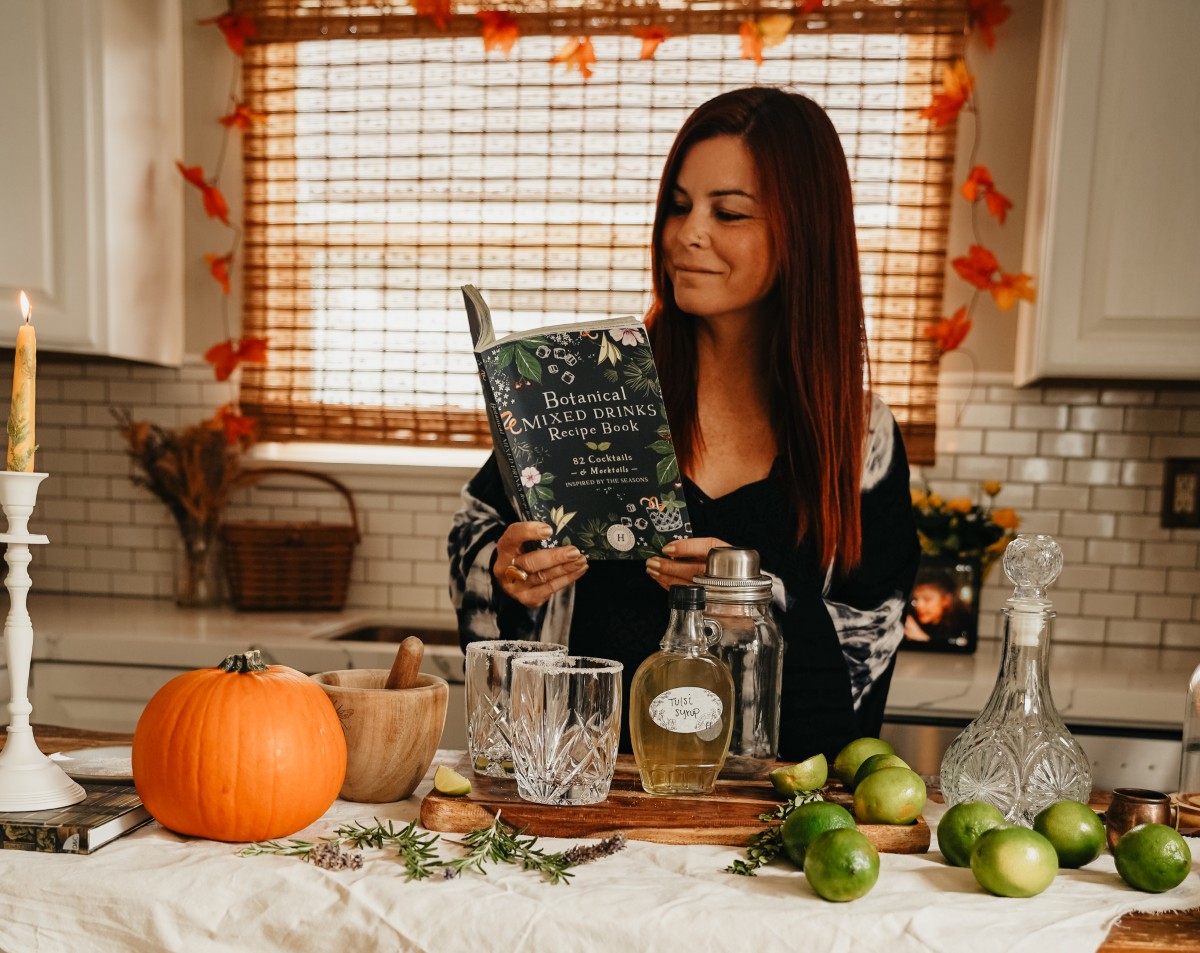 herbalist looking at Botanical Mixed Drinks Recipe Book with ingredients for Tulsi Lime Margarita on the counter