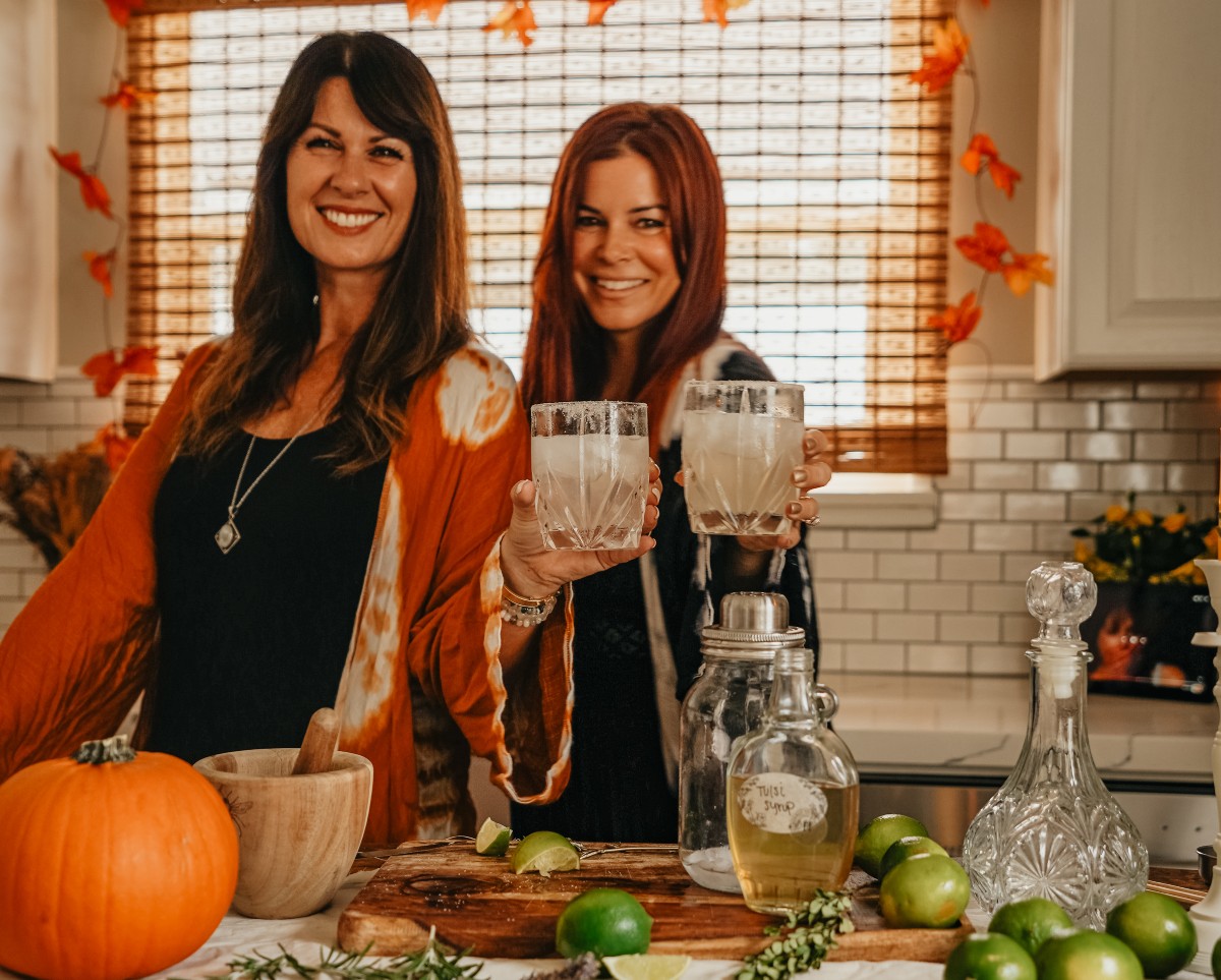 two herbalists smiling at the camera holding glasses of margarita toward the camera with bottles, limes, and a pumpkin on counter